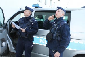 policjanci na tle radiowozu spoglądający w górę. Jedne trzyma w dłoni dokument drugi rozmawia przez telefon