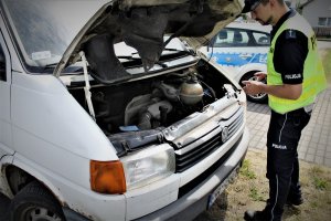 Policjant Wydziału Ruchu Drogowego dokonuje kontroli stanu technicznego pojazdu. Pochyla się nad komorą silnika kontrolowanego pojazdu typu bus.