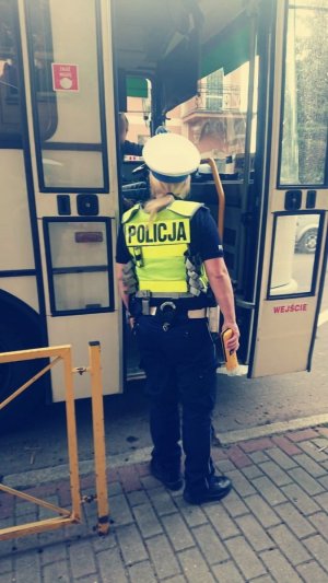 Policjanta z Wydziału Ruchu Drogowego w kamizelce odblaskowej z napisem Policja stojąca tyłem przy otwartych drzwiach autokaru szkolnego. Na zdjęciu widać jedynie fragment autobusu z frontowymi drzwiami.
