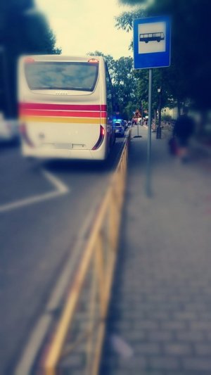Autobus szkolny koloru białego stojący na przystanku autobusowym- widać niebieski znak oznaczający przystanek autobusowy oraz fragment chodnika. Zdjęcie częściowo rozmyte ostrość skupiona na autokarze.