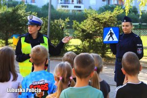 Policjanta z wydziału duchu drogowego wskazuje rękoma na znaj oznaczający przejście dla pieszych. Druga policjanta trzyma ten znak w sposób widoczny na dzieci, które słuchają policjantki. Na zdjęciu widać dzieci ustawione przodem do Policjantek