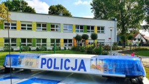 napis policja z gondoli radiowozu w tle placówka oświatowa oraz dzieci i policjanci wychodzący ze placówki.