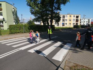 Policjanci i dzieci na oznakowanym przejściu dla pieszych. Policjanci uczą prawidłowych zachowań w trakcie przekraczania jezdni. Dzieci na pasach ubrane w kamizelki odblaskowe