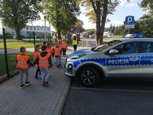 dzieci ubrane w kamizelki odblaskowe dwójkami kierują się za policjantką w kierunku przejścia dla pieszych. Po prawej stronie widać oznakowany radiowóz policyjny.