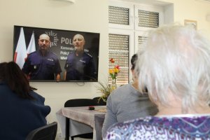 Seniorzy ustawieni tyłem do zdjęcia oglądający ekran telewizora na którym widać dwóch umundurowanych policjantów