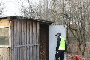 policjant kontrolujący drewnianą altanę pod kątem przebywania osób bezdomnych. Policjant stoi przy drzwiach altany i patrzy do środka