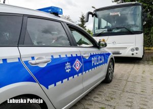 bok radiowozu oznakowanego z napisem POLICJA na tle przedniej części autobusu