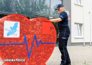 policjantka stojąca przy metalowym  charytatywnym sercu w trakcie wrzucania nakrętek
