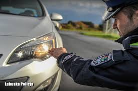 Policjant wskazujący lewą dłonią w reflektor pojazdu w którym włączone są światła mijania