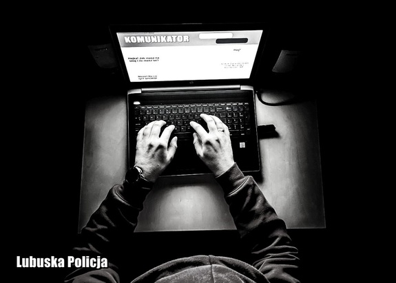 zdjęcie czarno-białe. Widok z góry na posturę człowieka w kapturze trzymającego dłonie na klawiaturze przed ekranem komputera