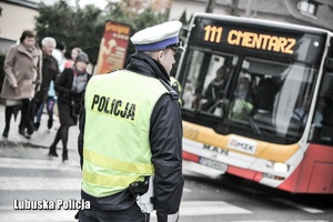 policjant stojący przy oznakowanym przejściu dla pieszych  w tle autokar z napisem: Cmentarz.