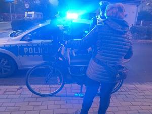 na zdjęciu widzimy policjantów stojących przy radiowozie z włączonymi sygnałami świetlnymi, przeprowadzający kontrolę drogową wobec osoby poruszającej się rowerem.