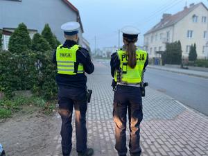 na zdjęciu widzimy dwóch funkcjonariuszy wydziału ruchu drogowego stojących  tyłem ubranych w żółte kamizelki odblaskowe